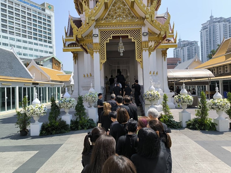 The outside of a temple for funeral services in Thailand with a queue of mourners in black.