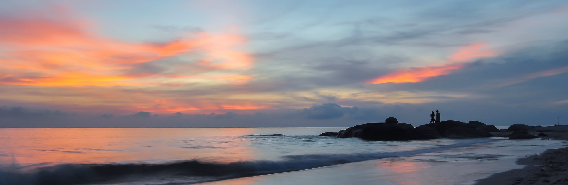 Sunset over a sandy beach on Thailand's Gulf coast, with two people standing on the rocks